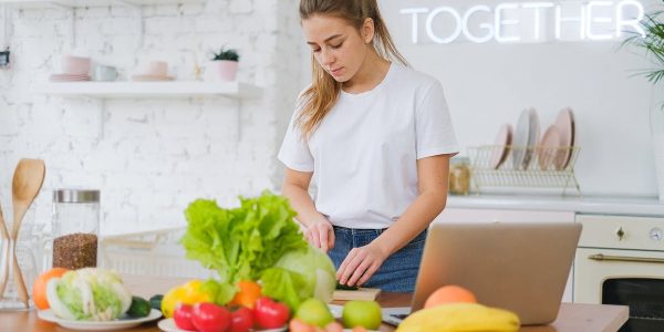 Woman in white t-shirt preparing fresh vegetables in a bright kitchen with a laptop.